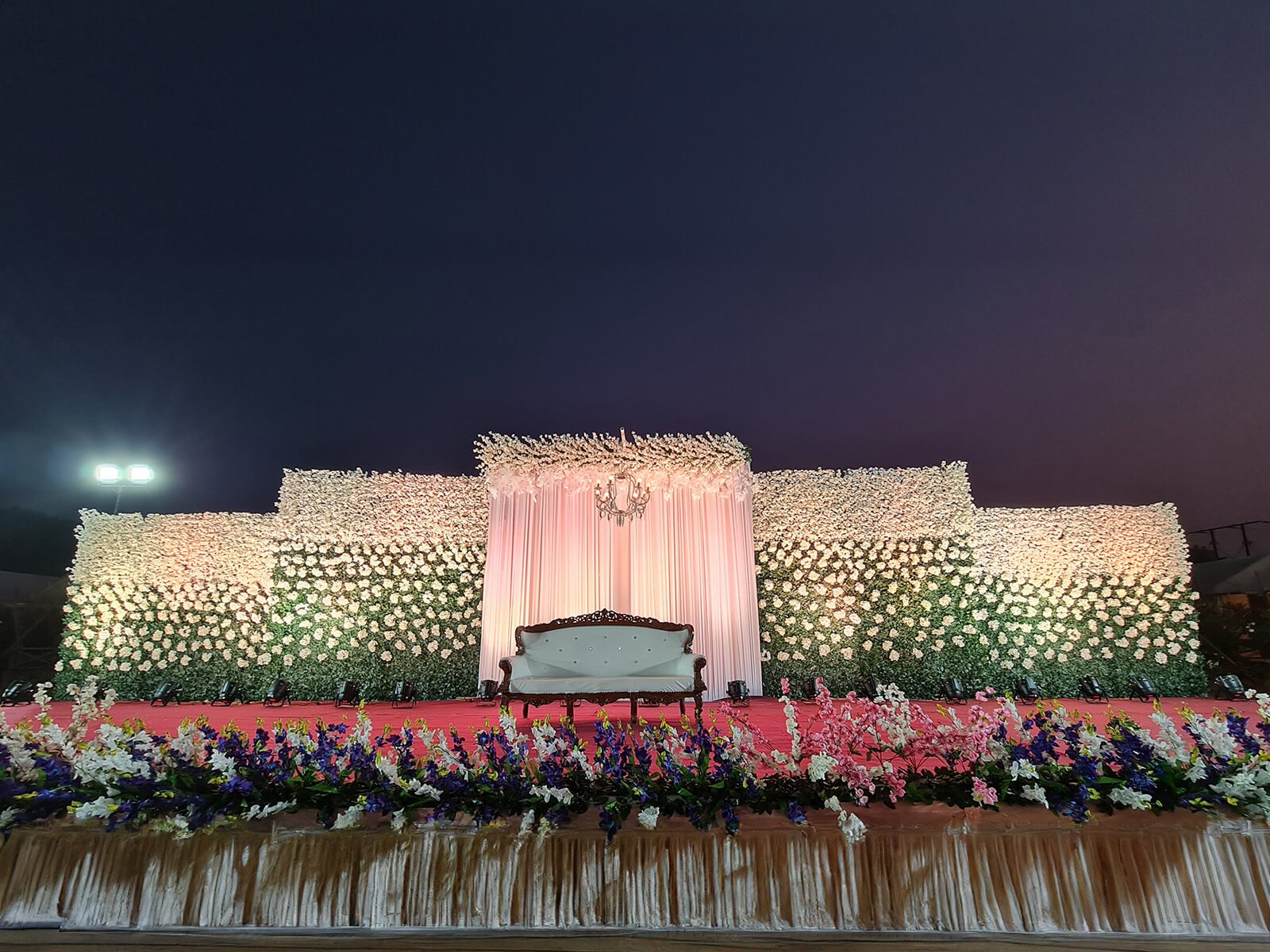 Outdoor wedding stage with a backdrop adorned with white flowers, a central pink draped section, a chandelier, and a vintage-style white sofa, with a foreground of colorful floral arrangements.