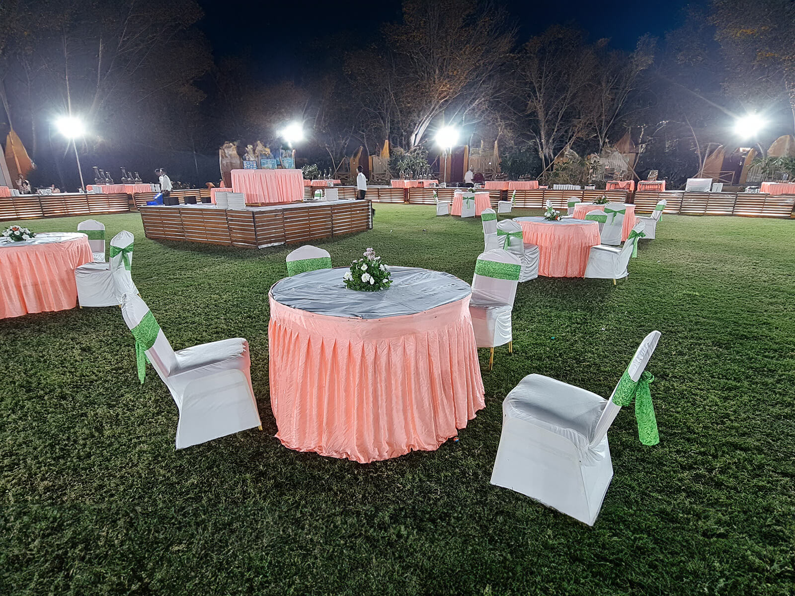 Outdoor evening event with tables and chairs covered in white and pink tablecloths, green chair sashes, and floral centerpieces, illuminated by bright lights.