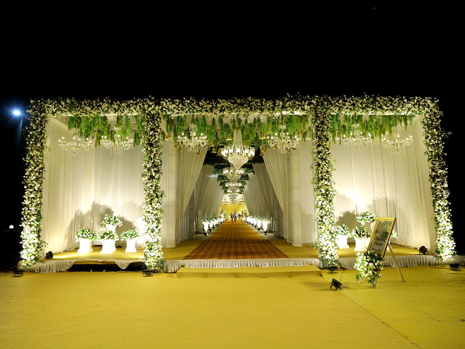 Grand wedding entrance decorated with white flowers, green foliage, chandeliers, and white drapery at night.