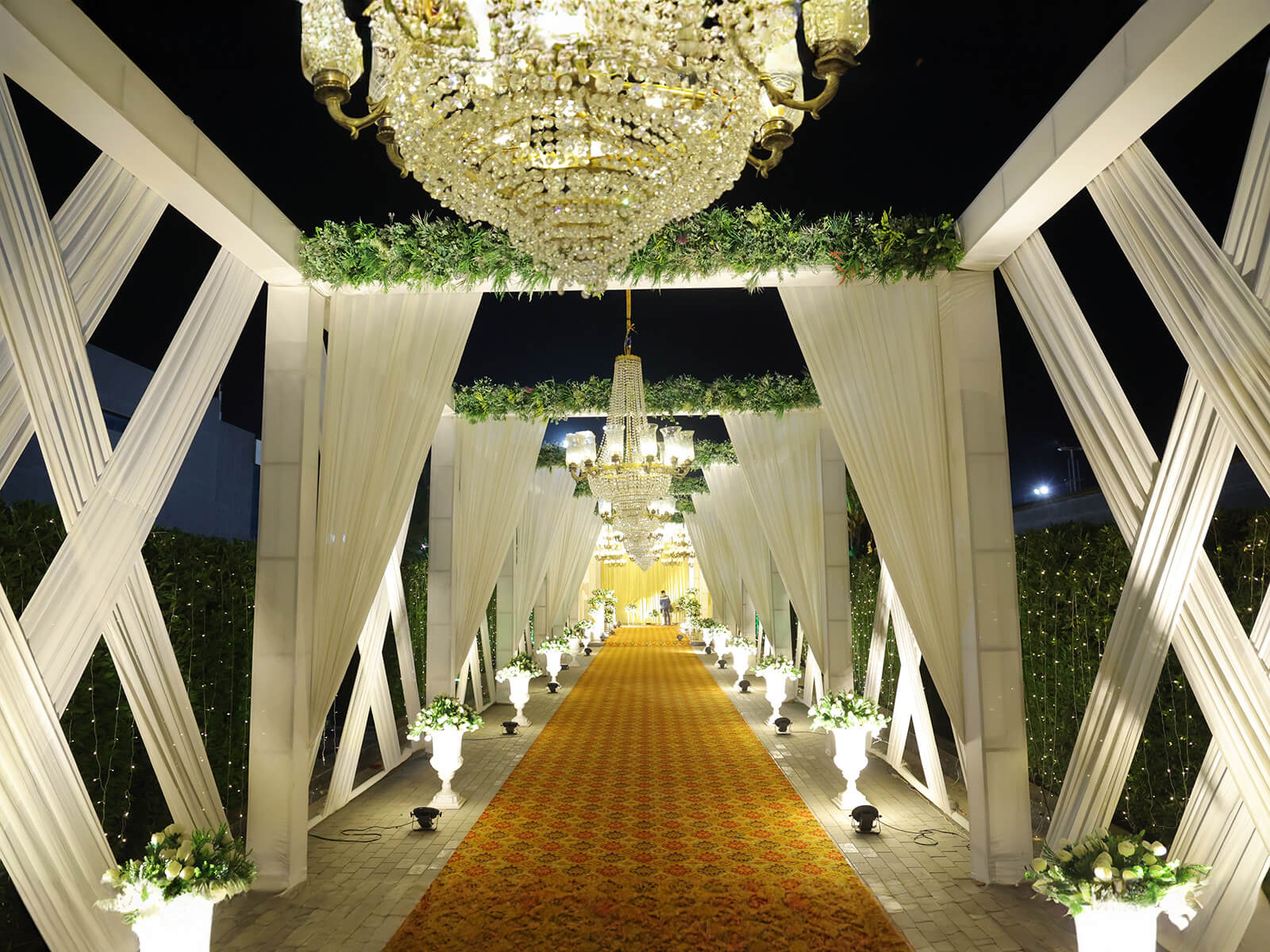 Elegant wedding entrance adorned with chandeliers, white drapery, and floral arrangements at night.