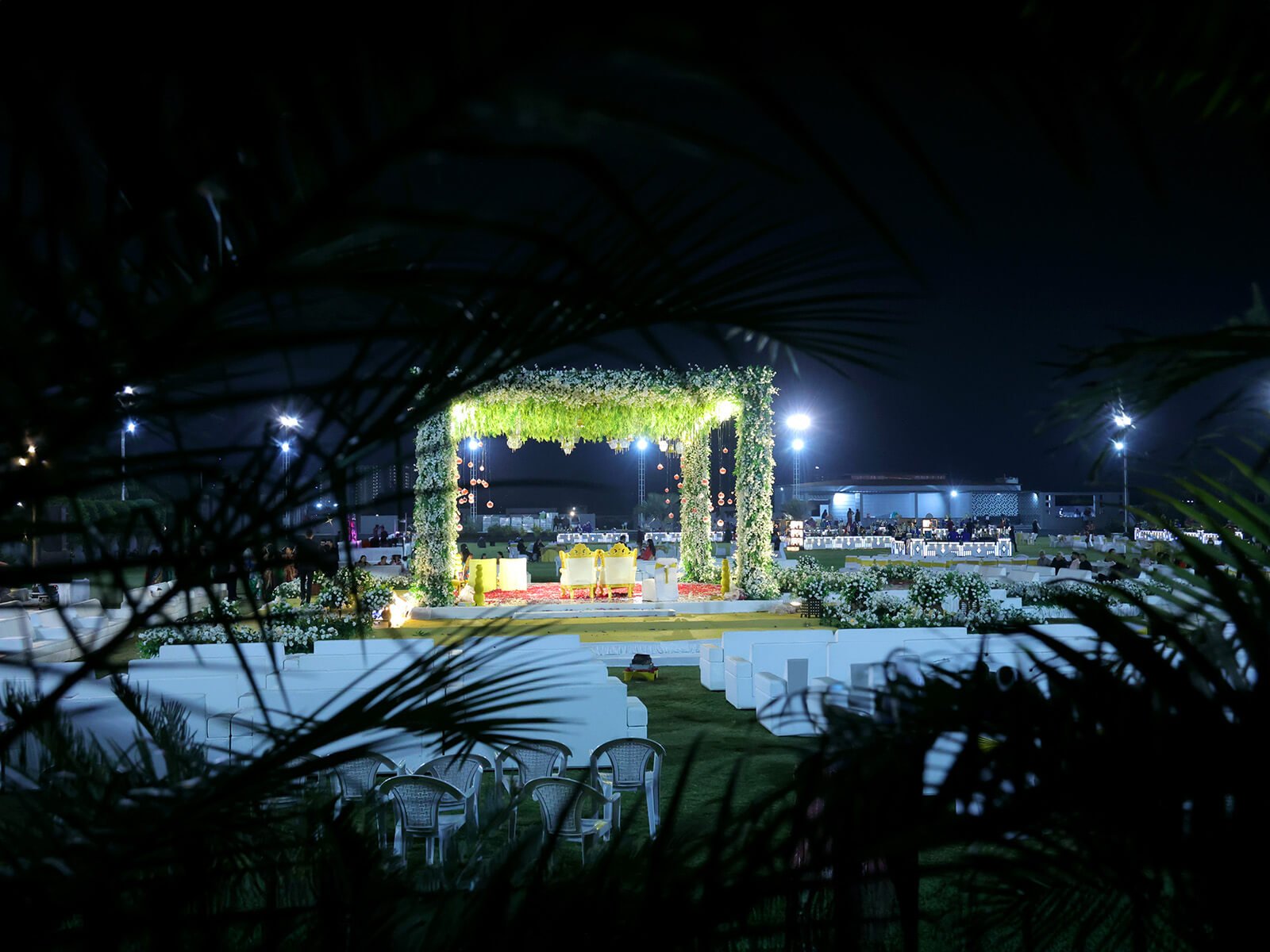 Nighttime outdoor wedding stage with greenery and lights, viewed through palm leaves.