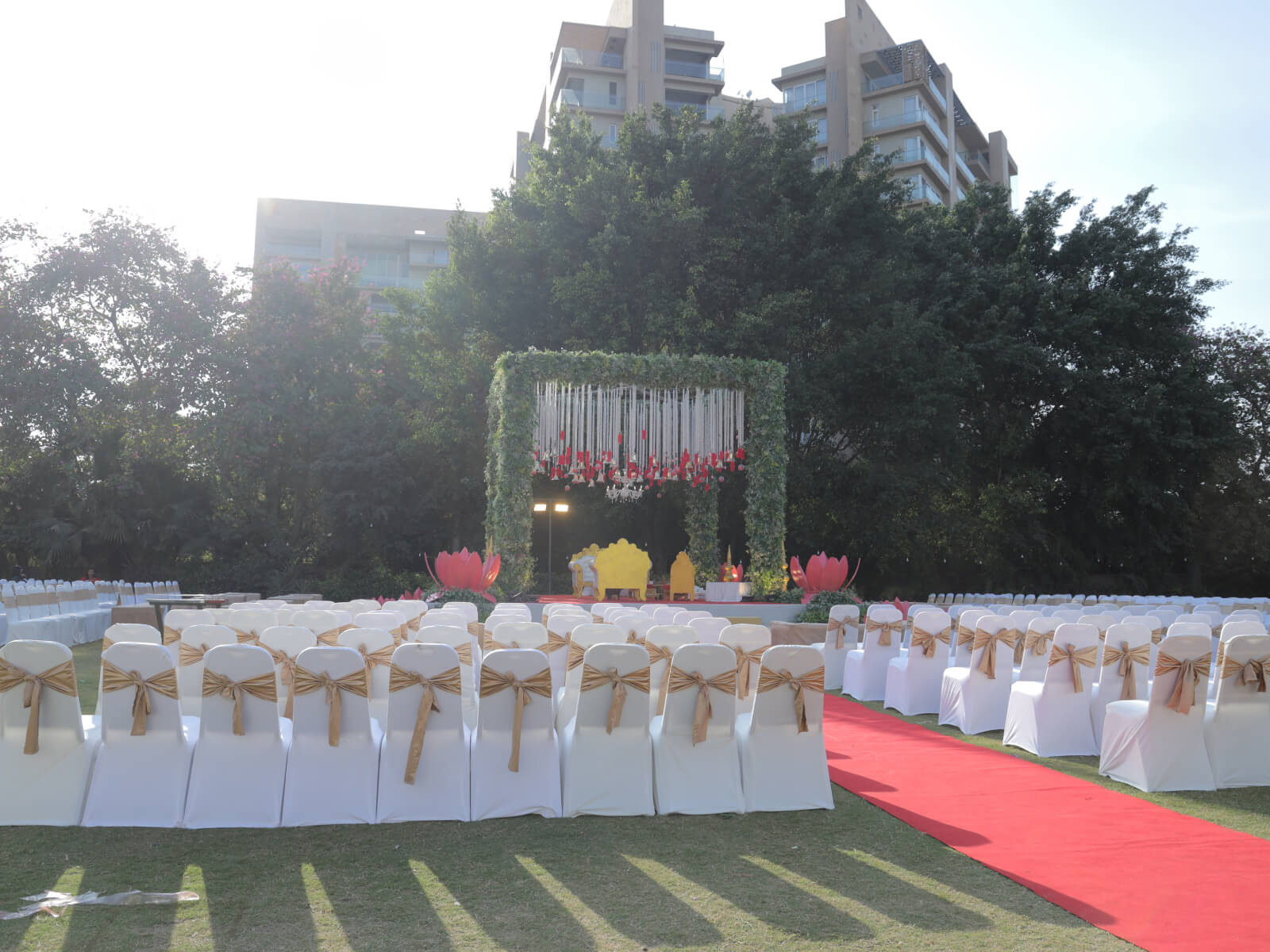 Outdoor wedding setup with white chairs and gold bows on a green lawn. The stage is decorated with greenery and hanging floral arrangements. Pink lotus elements add a touch of elegance. Urban buildings are visible in the background.