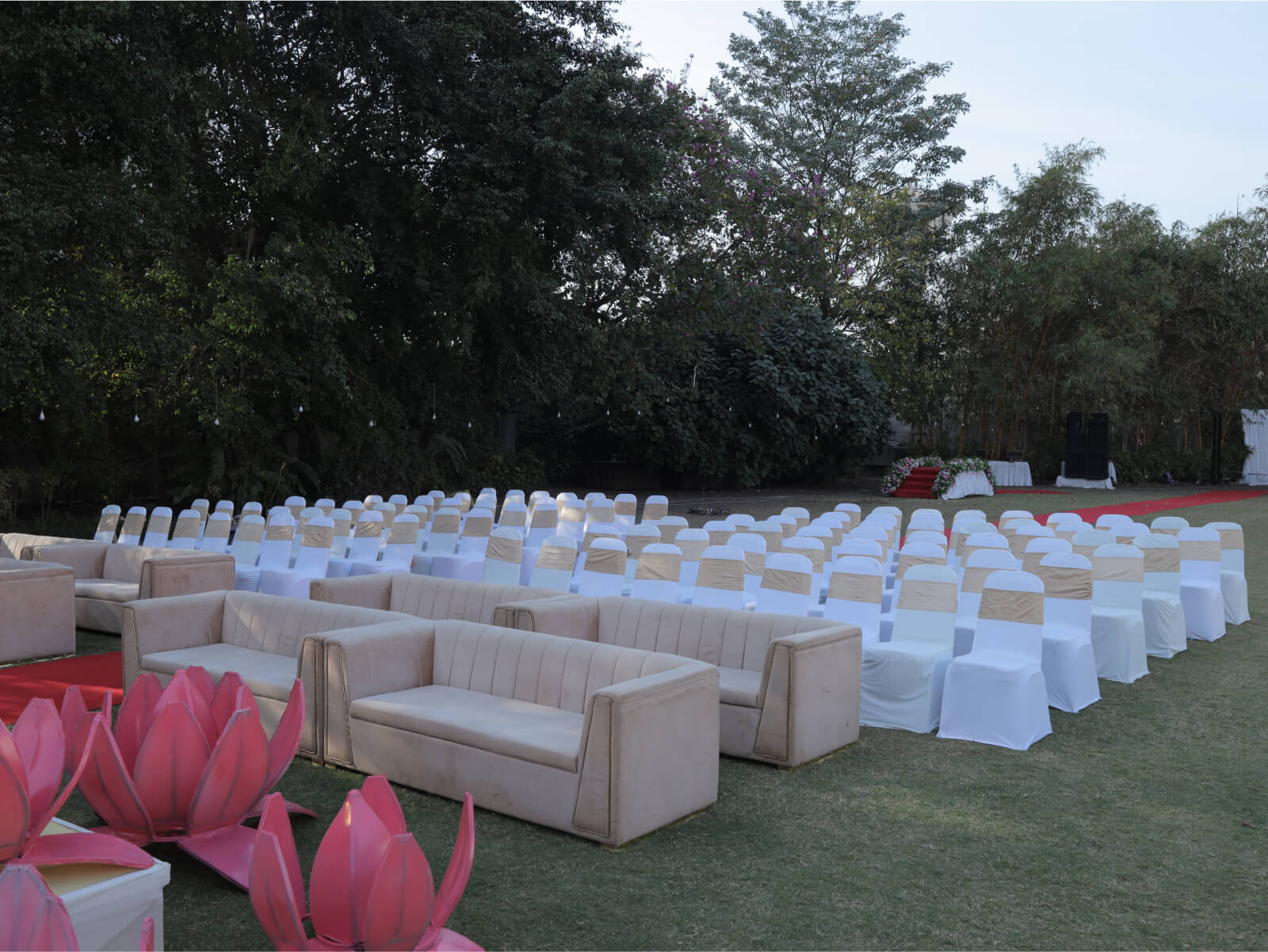 Outdoor wedding seating arrangement featuring white and gold-covered chairs and beige sofas, set in a lush garden with trees and a backdrop of greenery. Pink lotus-shaped decor elements enhance the elegant and serene atmosphere, perfect for a sophisticated event.