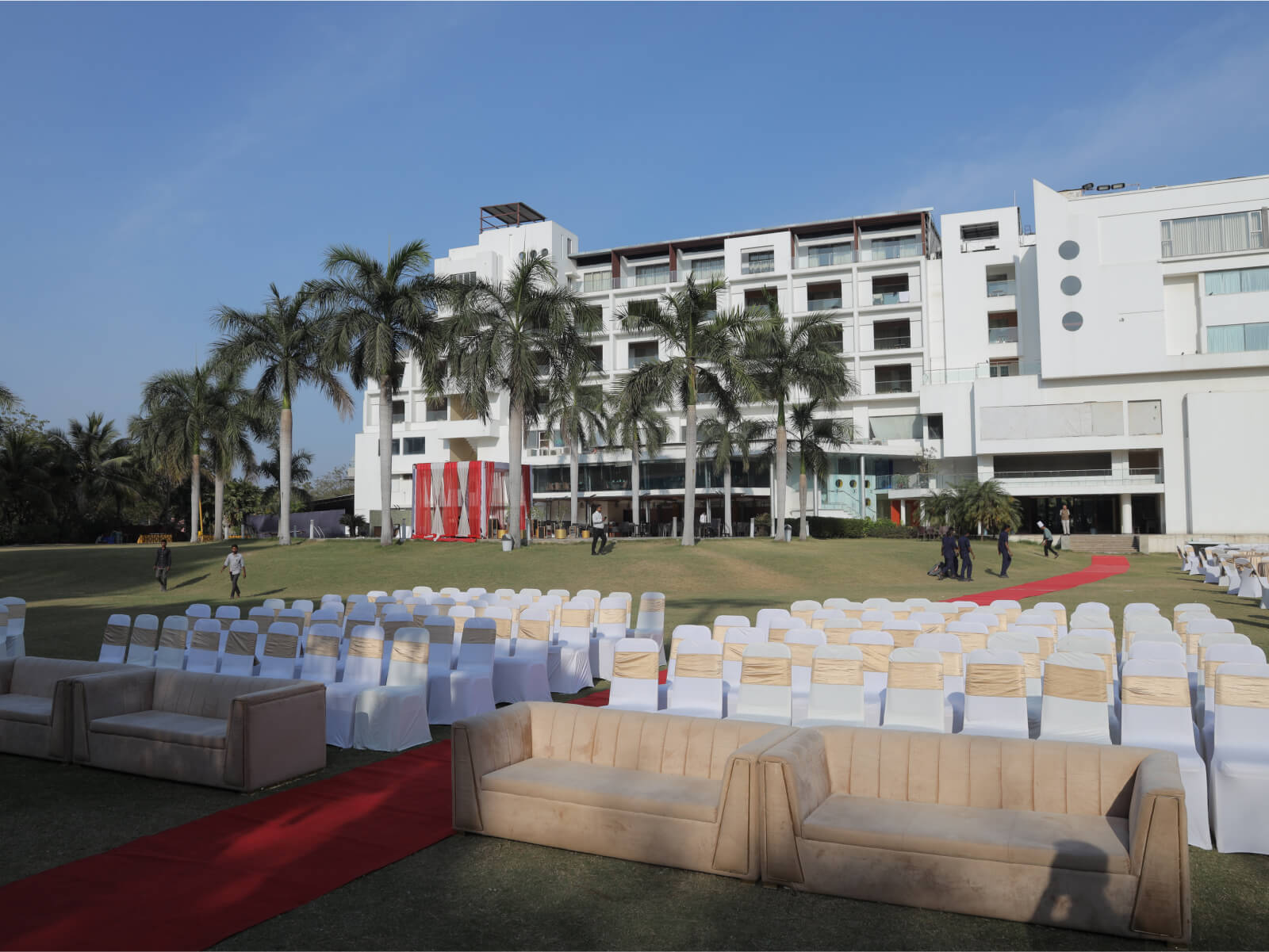 Outdoor wedding dining area with elegant white and gold-themed tables and chairs, set against the backdrop of a modern building and lush greenery. The setup includes food stalls labeled "CHAAT" and "SOUTH INDIAN," creating a festive and inviting atmosphere.