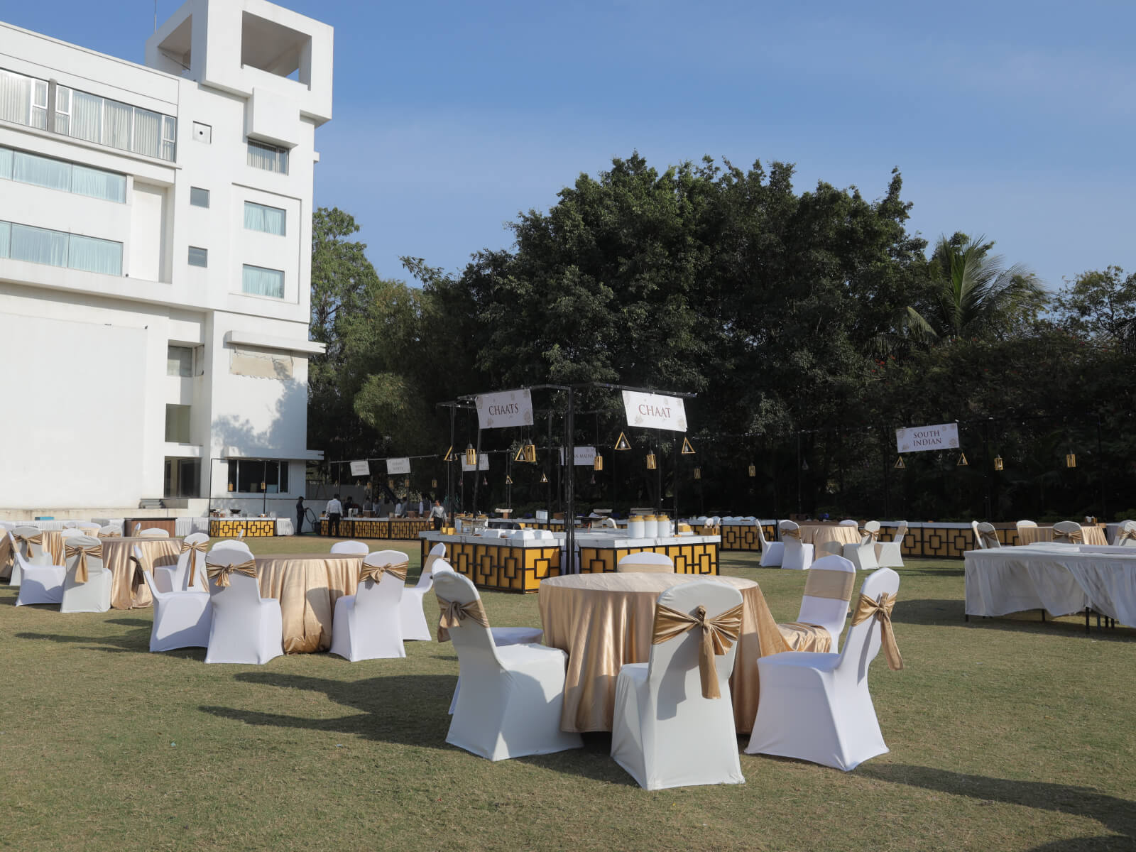 Outdoor wedding dining area with elegant white and gold-themed tables and chairs, set against the backdrop of a modern building and lush greenery. The setup includes food stalls labeled "CHAAT" and "SOUTH INDIAN," creating a festive and inviting atmosphere.