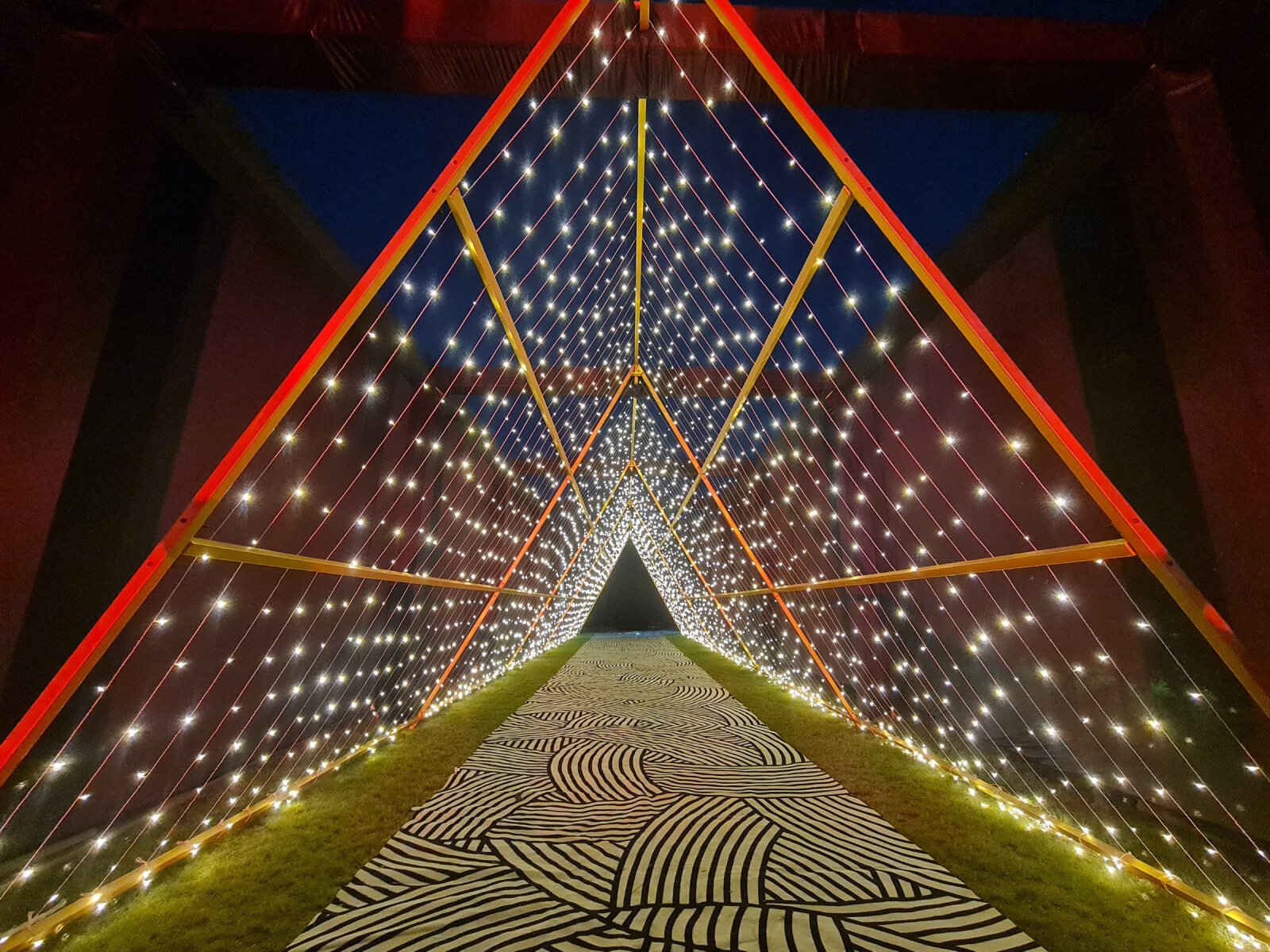 Indoor event entrance with illuminated triangular structure and decorative pathway, ideal for wedding decoration, event planning, and sangeet ceremonies.