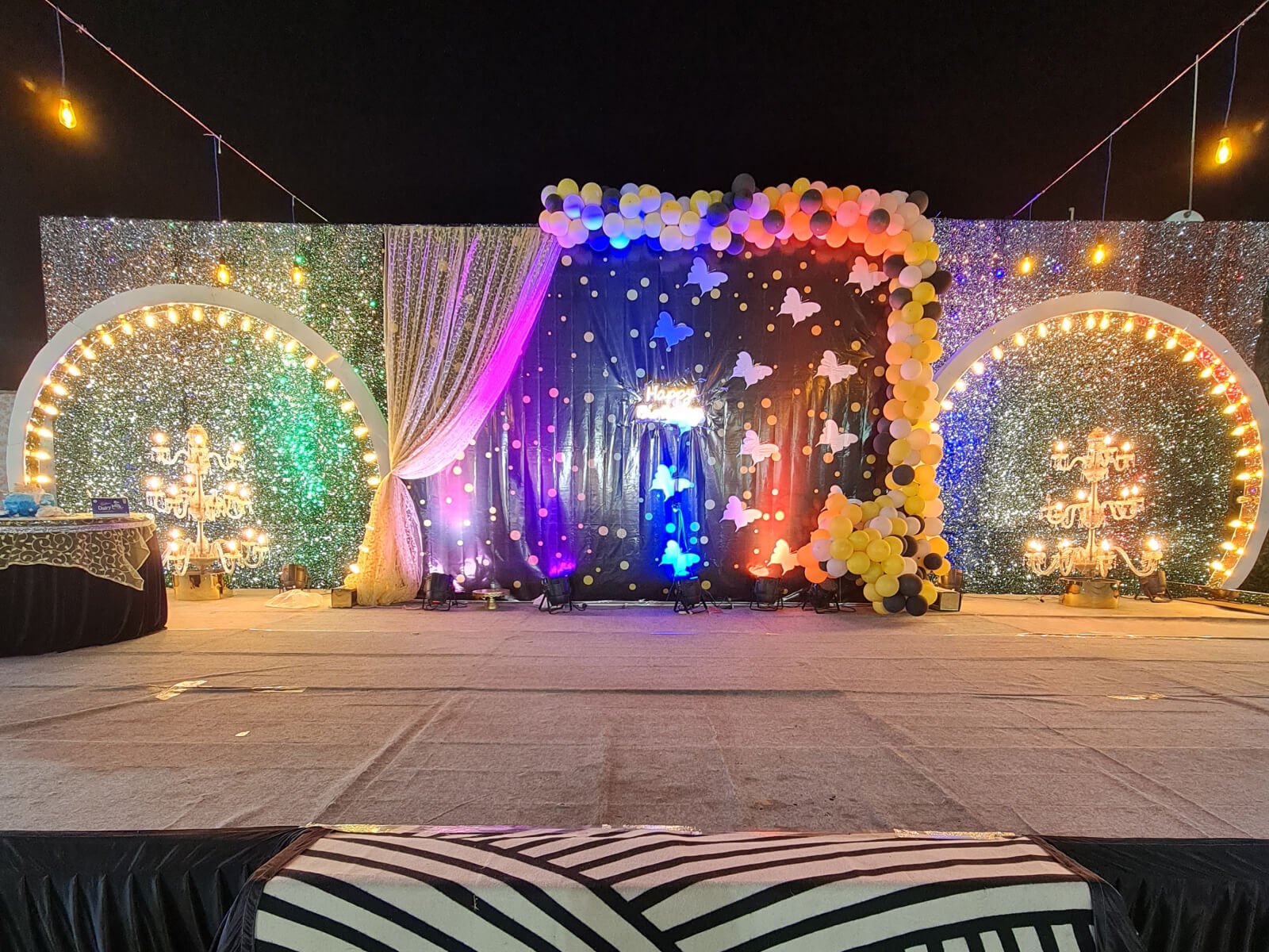 Outdoor event stage with balloon decorations, chandeliers, and illuminated backdrop.