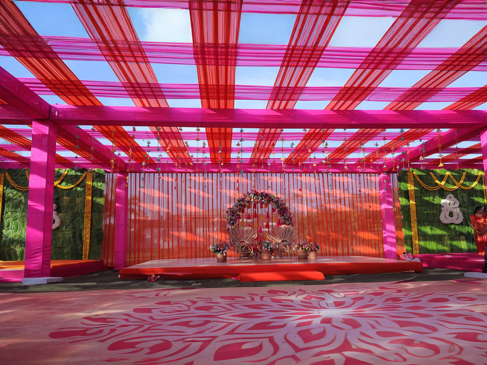 Outdoor wedding setup with vibrant pink and orange drapes, floral decorations, and colorful floor designs, ideal for wedding ceremonies and festive celebrations.