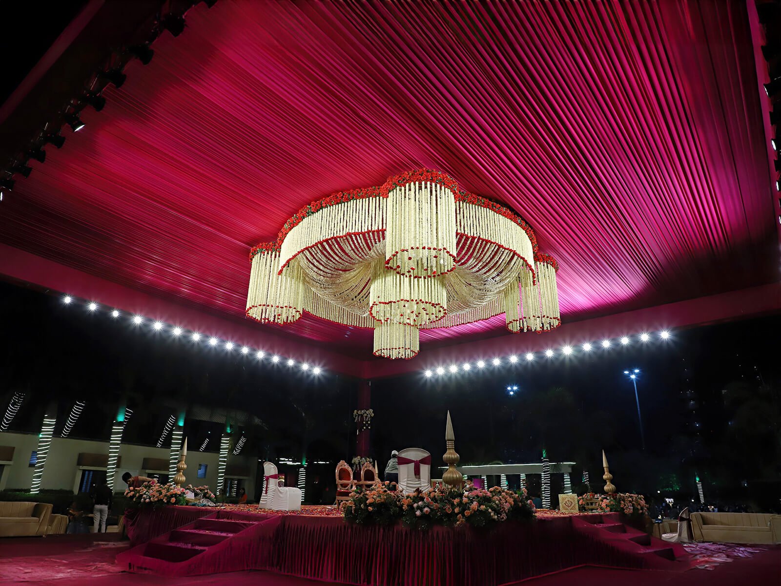 Outdoor wedding stage with vibrant drapes, floral decorations, and elegant chandelier lighting.