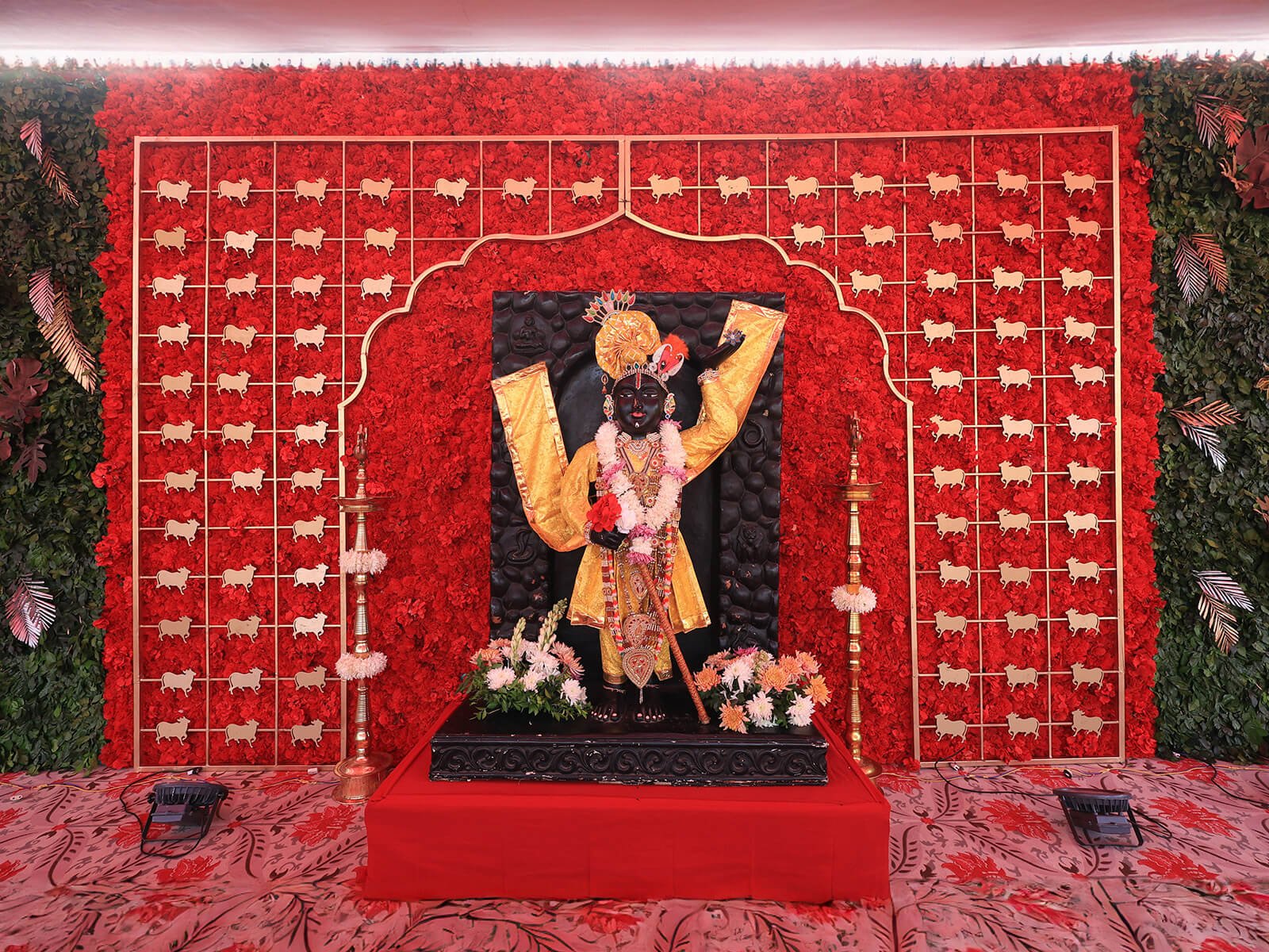 Indoor wedding decoration for a Haldi event featuring a Hindu deity statue with vibrant floral backdrop and traditional oil lamps.