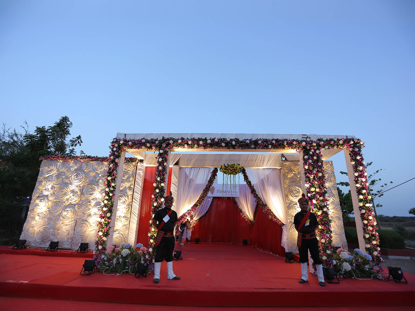 Outdoor wedding entrance with floral decorations, white drapes, and elegant lighting.