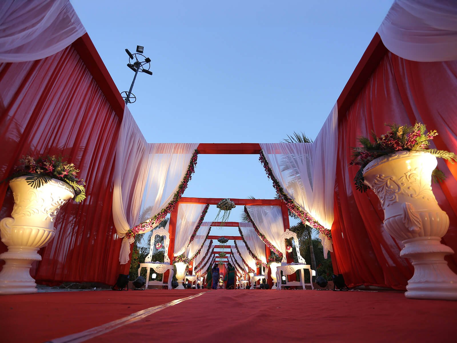 Outdoor wedding entrance with red drapes, floral arrangements, and elegant decor.