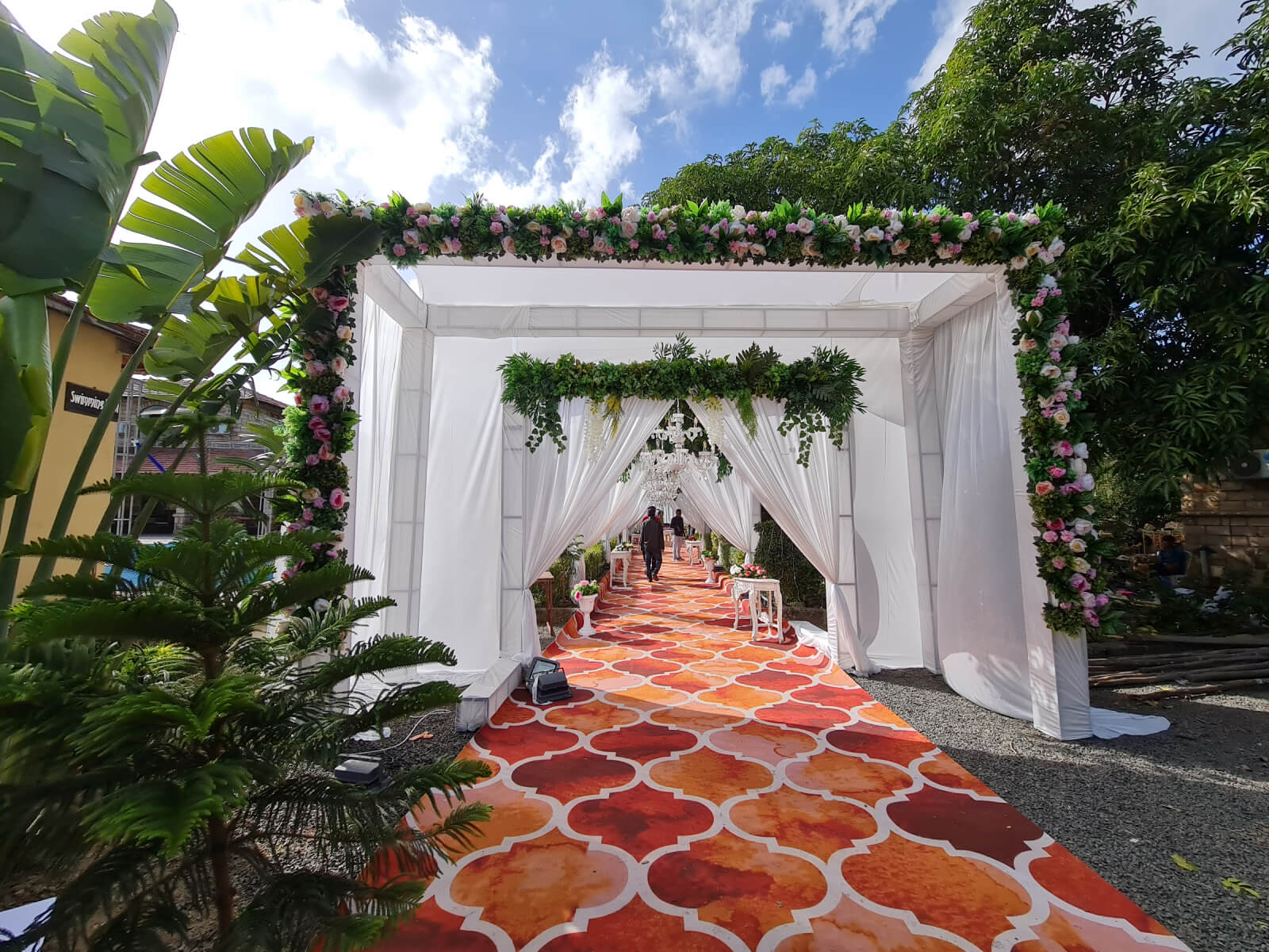 Beautifully decorated entrance with elegant white drapes and greenery, featuring a stylish chandelier and vibrant carpet, creating a grand and welcoming atmosphere for the wedding event.